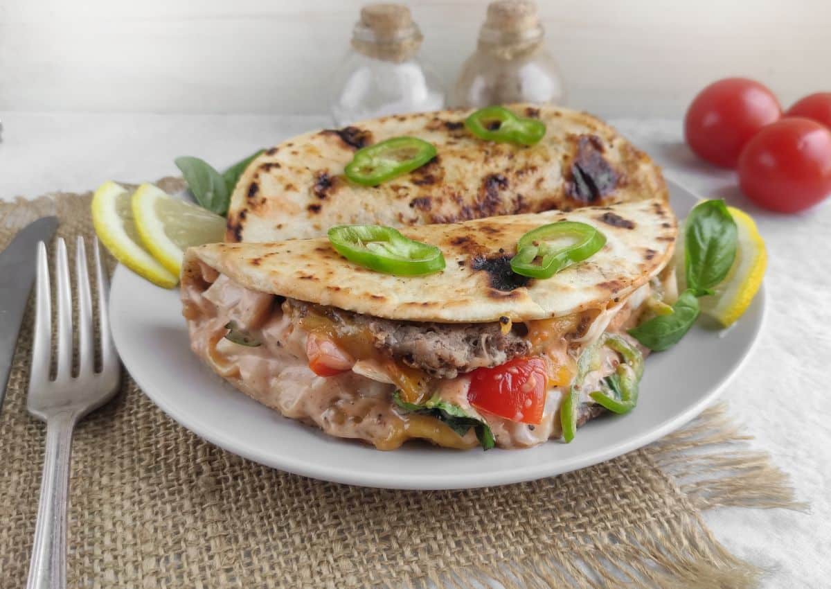 Two folded grilled tortillas filled with melted cheese, sliced tomatoes, greens, and topped with jalapeรฑo slices, served on a white plate with lemon wedges and fresh basil.