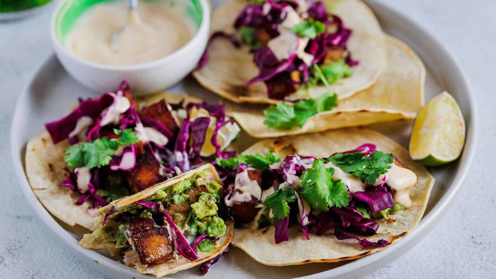 A plate with four tacos filled with diced vegetables, red cabbage, cilantro, sauce, and a lime wedge, with a bowl of creamy sauce on the side.
