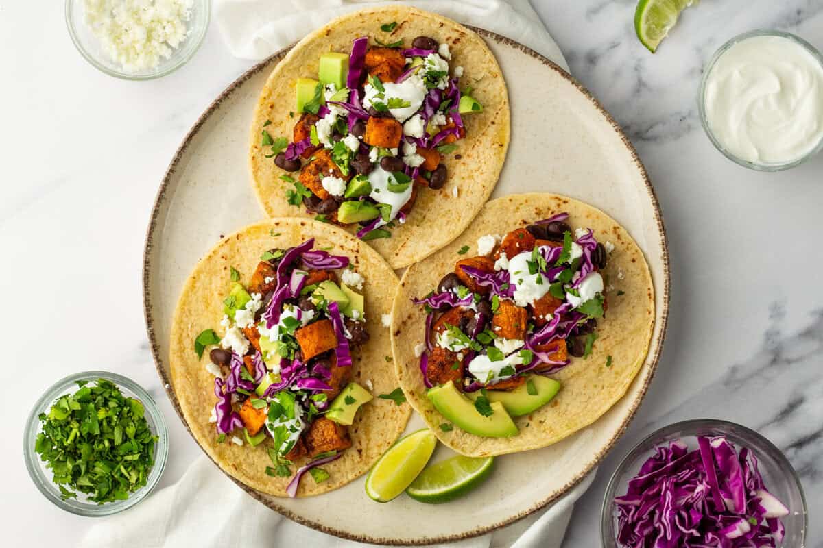 A plate with three tacos filled with roasted vegetables, avocado, black beans, purple cabbage, cilantro, and a drizzle of white sauce. Lime wedges and small bowls of toppings surround the plate.