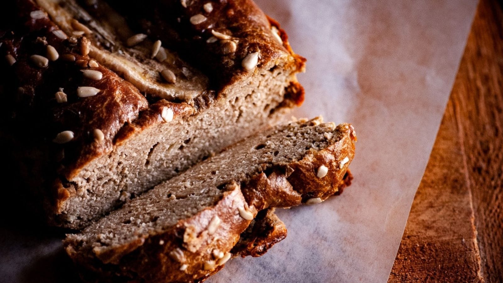 A loaf of banana bread topped with seeds sits on parchment paper, with one slice partially cut and resting in front of the loaf.