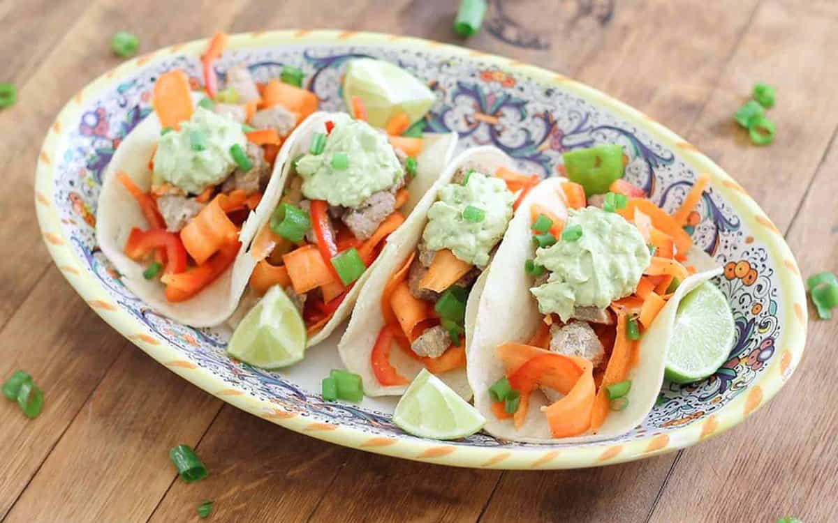 Three soft tacos filled with sliced vegetables, meat, and guacamole are arranged on a decorative plate with lime wedges and green onions on a wooden surface.