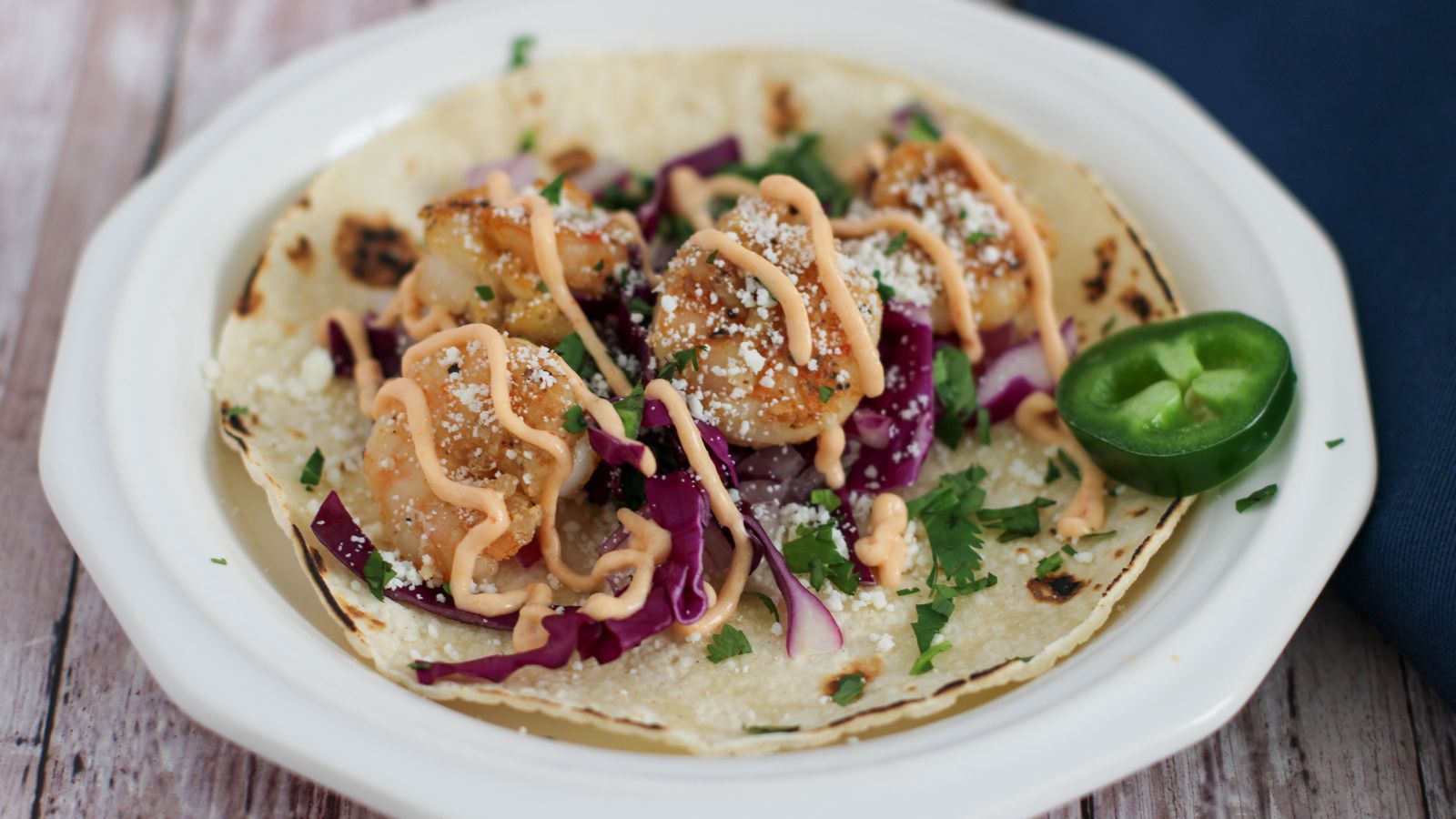 A taco on a white plate topped with breaded shrimp, purple cabbage, chopped cilantro, shredded cheese, a creamy sauce, and a sliced jalapeรฑo on the side.