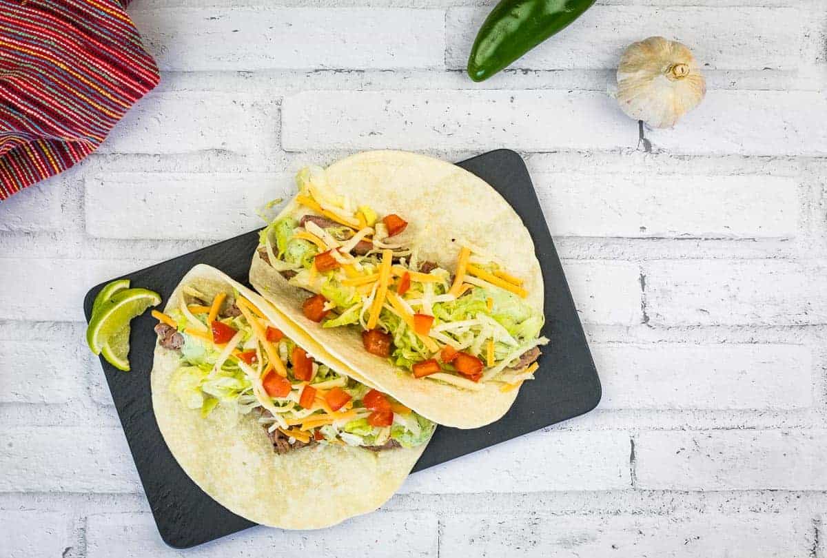 Two soft tacos filled with shredded lettuce, cheese, diced tomatoes, and meat, served on a black slate with lime wedges, on a white brick background.