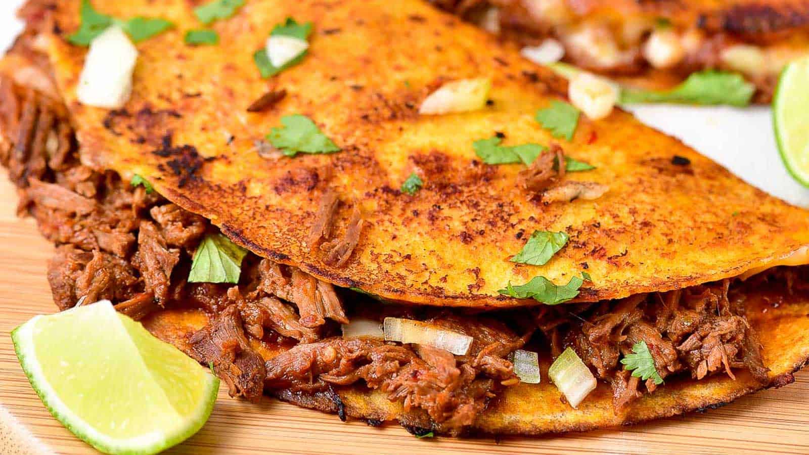 A close-up of a crispy taco filled with shredded beef, diced onions, and cilantro, served with a wedge of lime on a wooden surface.