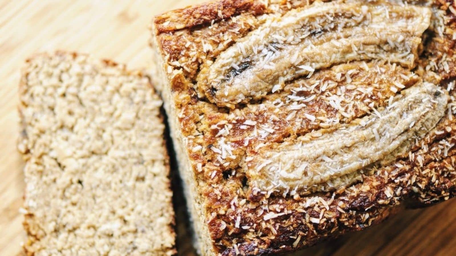 A loaf of banana bread topped with shredded coconut and banana slices, with one slice cut and resting beside it on a wooden surface.
