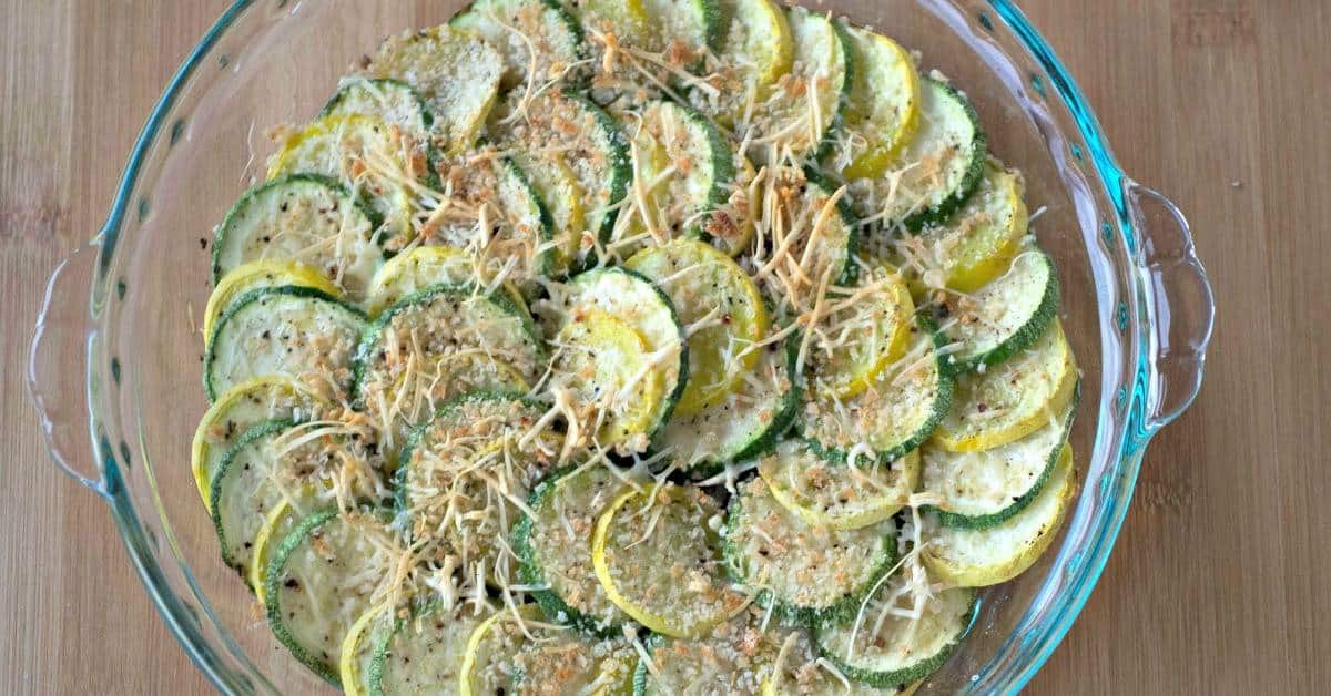 A glass dish filled with sliced yellow squash and zucchini, topped with shredded cheese and breadcrumbs, arranged in a circular pattern.