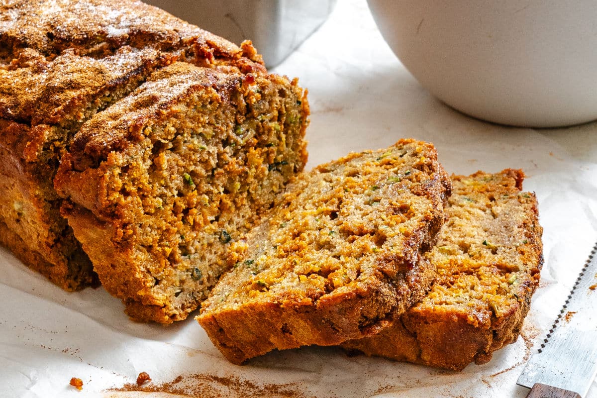 A sliced loaf of zucchini bread sits on parchment paper next to a bowl and a serrated knife.