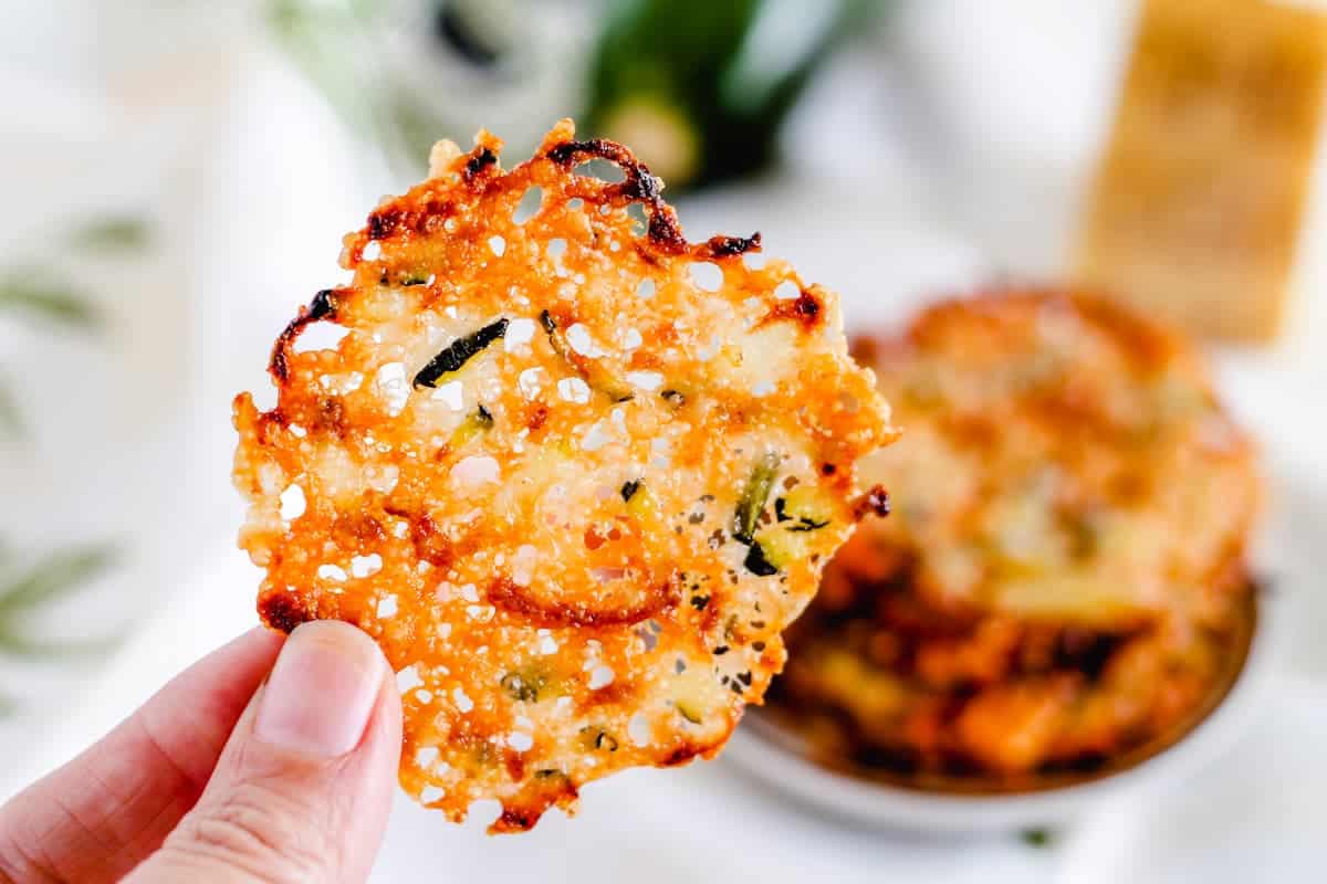 A hand holds a crispy, round parmesan cheese crisp with visible herbs and browned edges; a bowl with more crisps is blurred in the background.