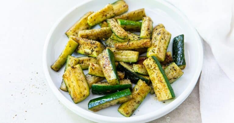 A white plate with seasoned, roasted zucchini sticks arranged in a single layer on a light surface.