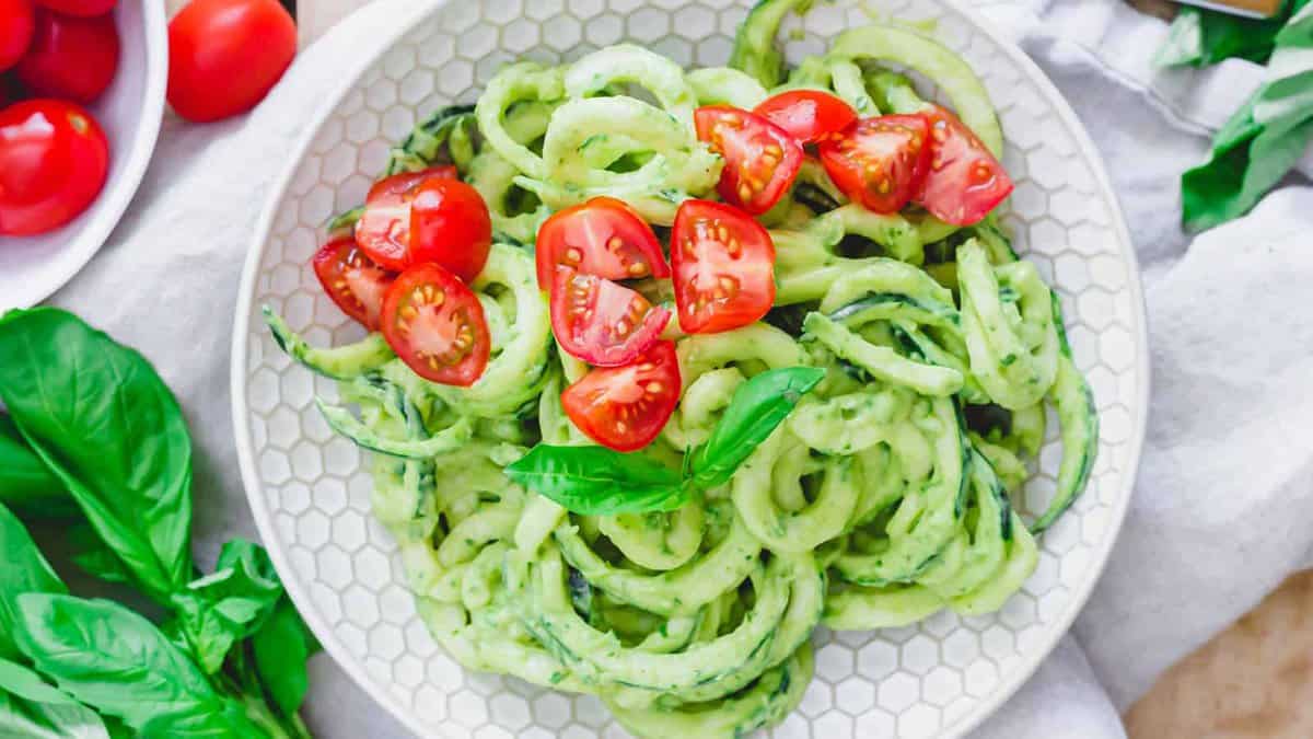 A plate of zucchini noodles topped with cherry tomato halves and garnished with a basil leaf, surrounded by fresh basil and tomatoes.