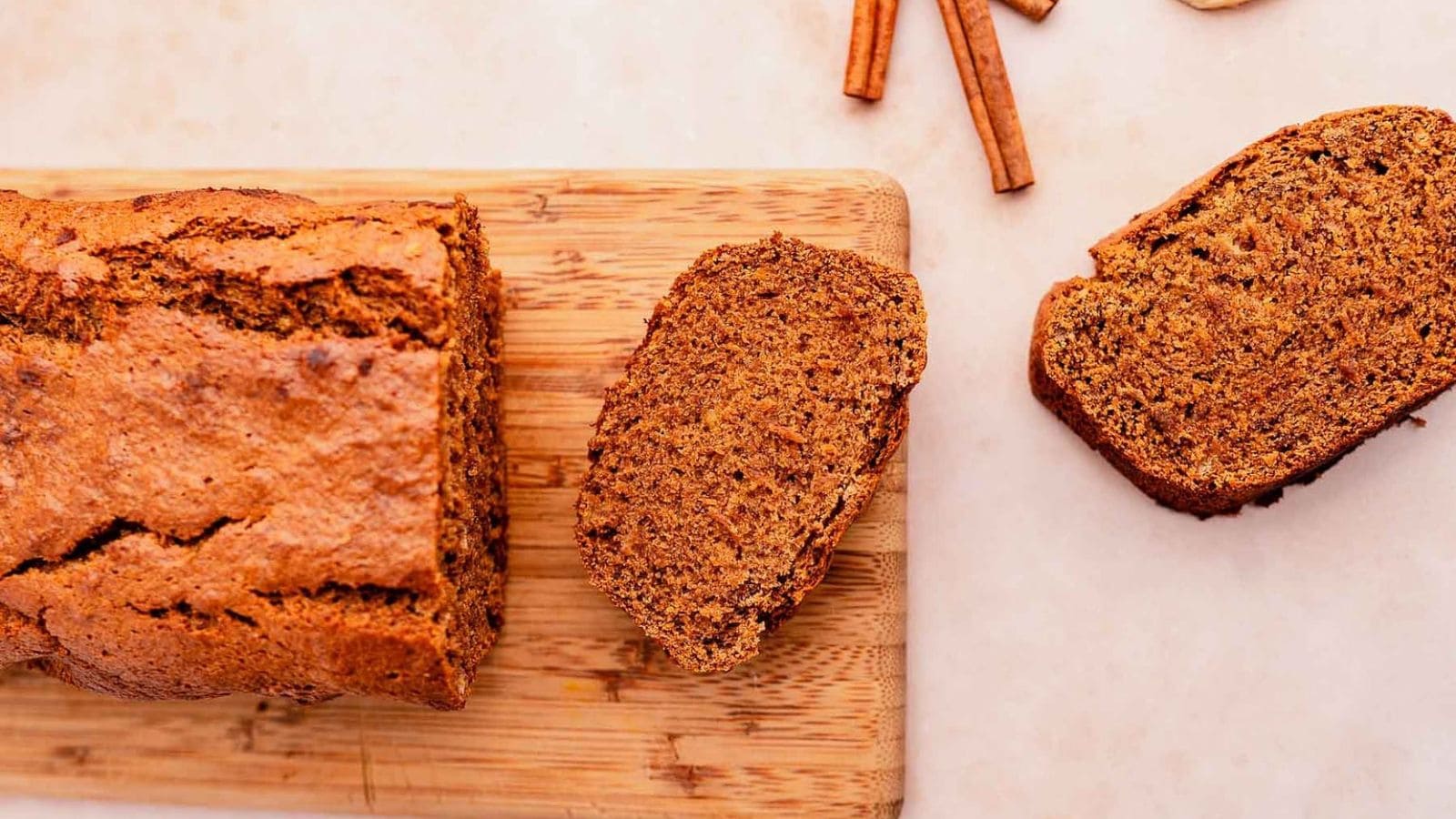 A loaf of sliced brown bread sits on a wooden cutting board, with two separate slices and cinnamon sticks beside it on a light surface.