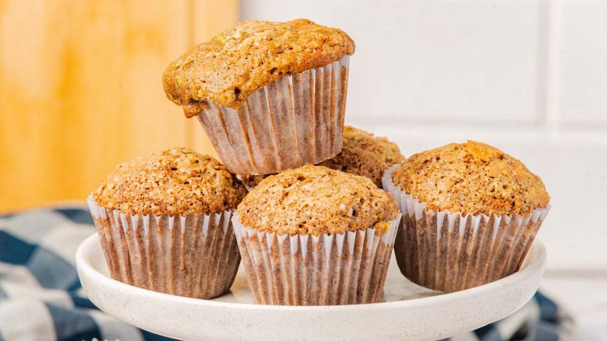 Five bran muffins in paper liners are arranged on a white cake stand, with one muffin resting on top of the others.