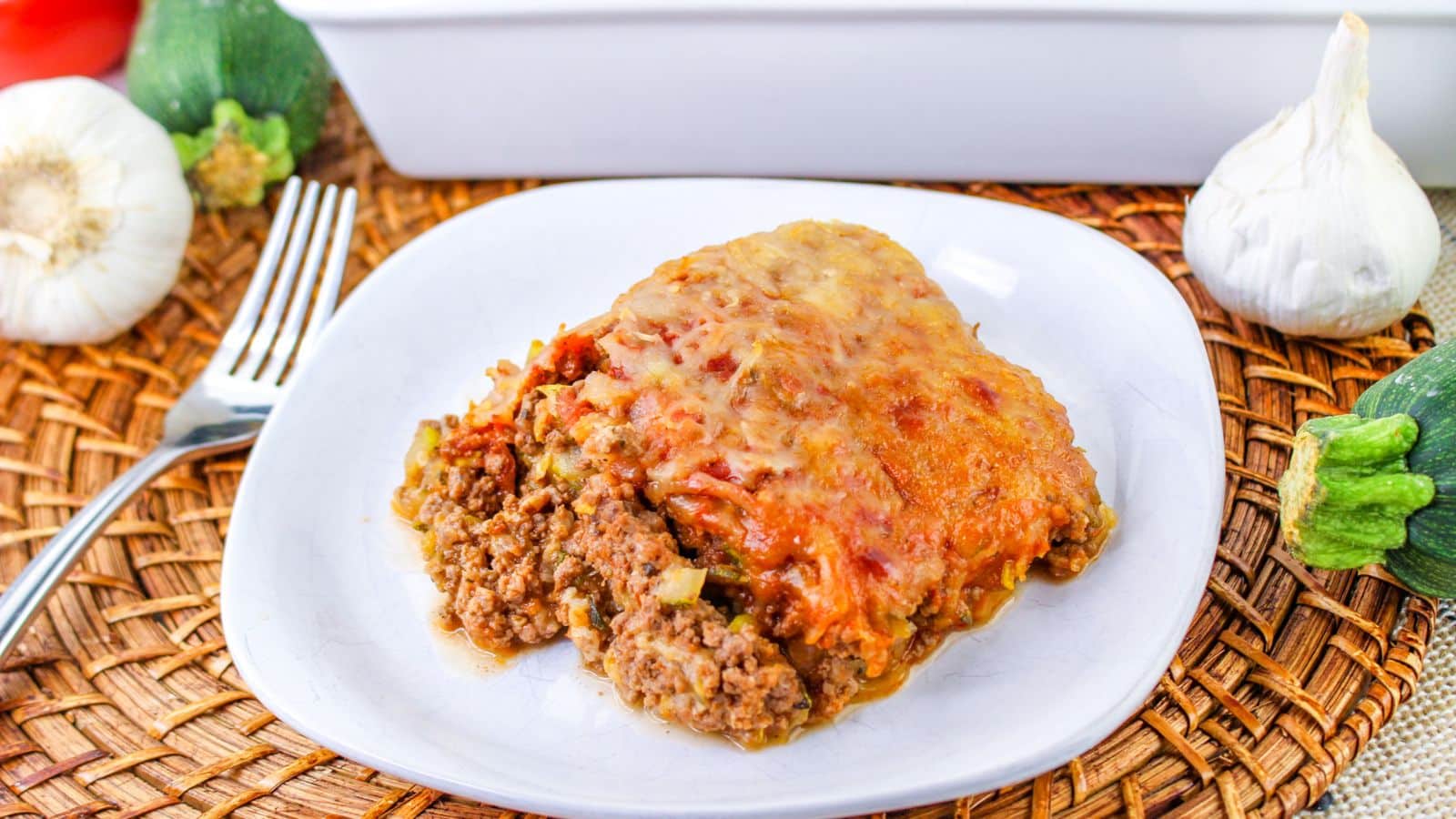 A slice of baked casserole with ground meat and melted cheese on a white plate, placed on a woven placemat with vegetables and a fork nearby.