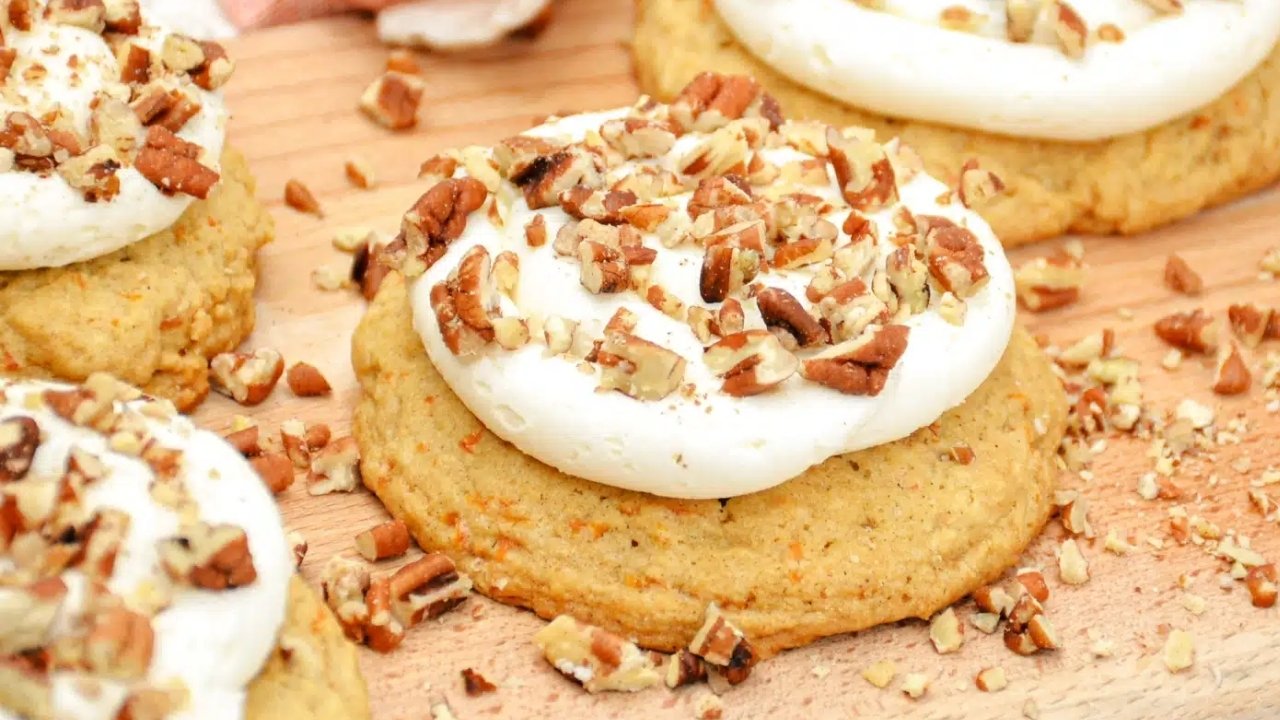 A close-up of cookies topped with white frosting and chopped pecans, arranged on a wooden surface.