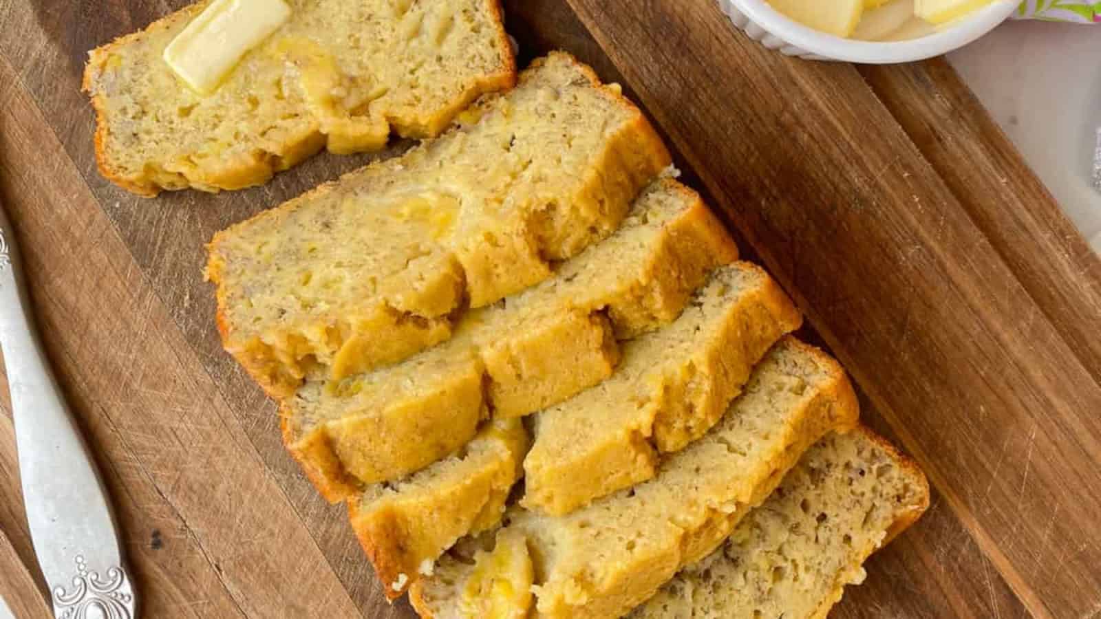 Several slices of banana bread are arranged on a wooden cutting board; one slice has a pat of butter on top.