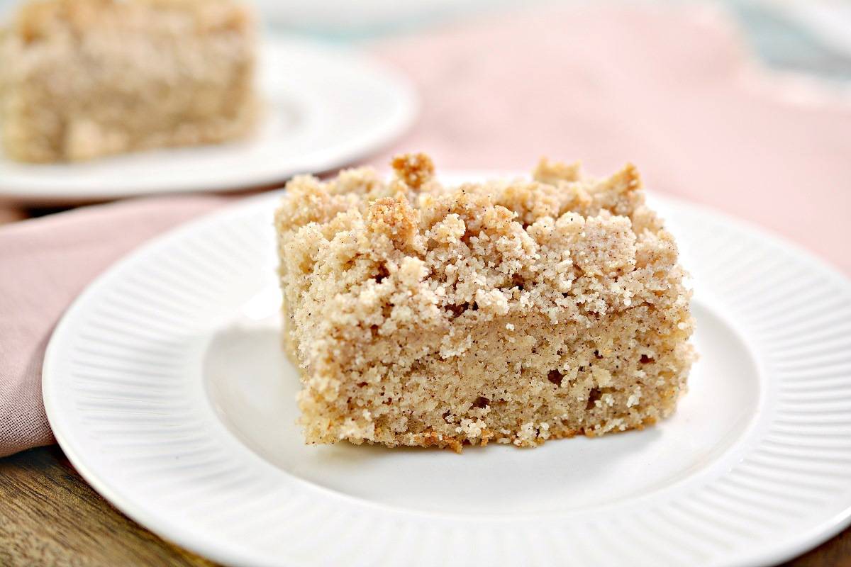 A square slice of crumb-topped coffee cake sits on a white plate, with a pink napkin and another plate in the background.