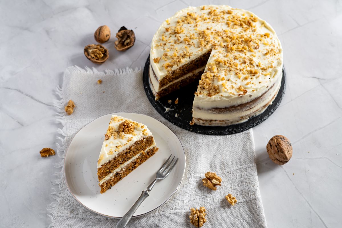 A round carrot cake with cream cheese frosting and walnut pieces sits on a tray, with one slice served on a plate beside it; whole walnuts are scattered around.