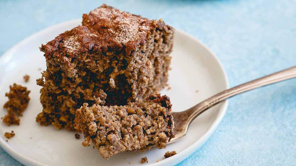 A piece of brown, crumbly cake sits on a white plate with a fork holding a bite-sized portion.