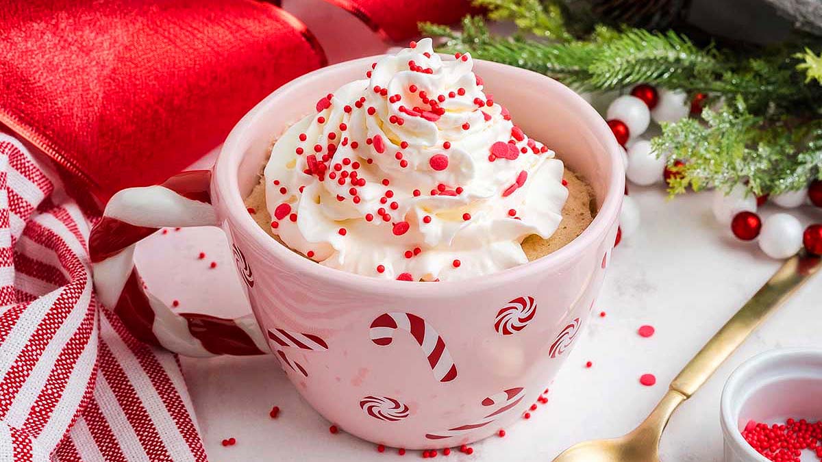 A pink mug with candy cane designs filled with a drink topped with whipped cream and red sprinkles, next to festive decor and a gold spoon.