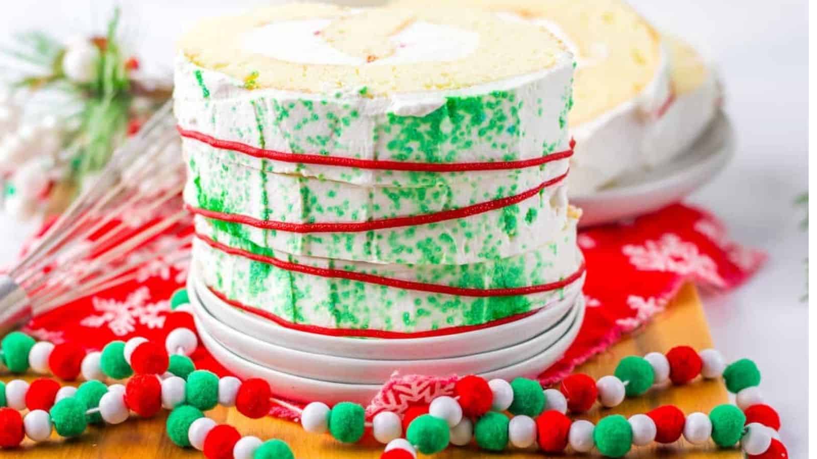 A festive cake roll with white frosting, green sprinkles, and red icing sits on a plate, surrounded by Christmas-themed decorations and colorful pom-pom garland.