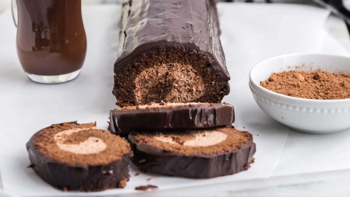 A chocolate Swiss roll cake with a cream filling is sliced on a white surface, next to a bowl of cocoa powder and a glass of chocolate drink.