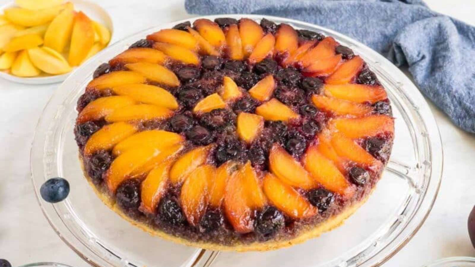 A round upside-down cake topped with arranged slices of peaches and whole blueberries sits on a glass cake stand. A bowl of sliced peaches is in the background.