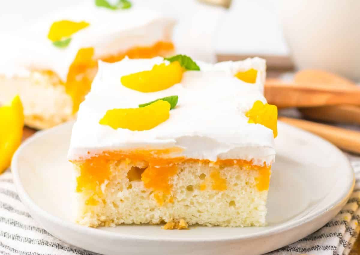 A slice of white cake topped with whipped cream and peach slices sits on a white plate.