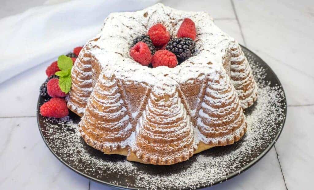 A bundt cake dusted with powdered sugar, topped with raspberries and blackberries, and garnished with mint leaves on a black plate.