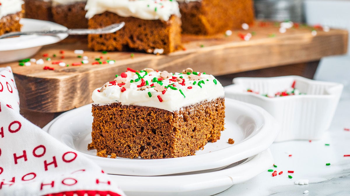 A square piece of gingerbread cake with white frosting and red, green, and white sprinkles sits on a white plate, with more cake pieces in the background.