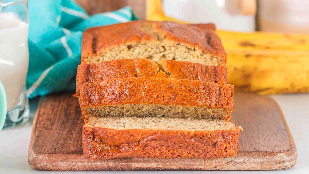 A loaf of banana bread with two slices cut, placed on a wooden cutting board. Bananas and a glass of milk are in the background.
