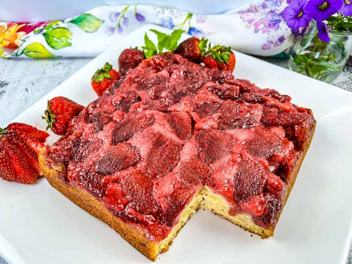 A square strawberry upside-down cake with a slice missing, topped with baked strawberries, is presented on a white plate. Fresh strawberries and a floral cloth are in the background.