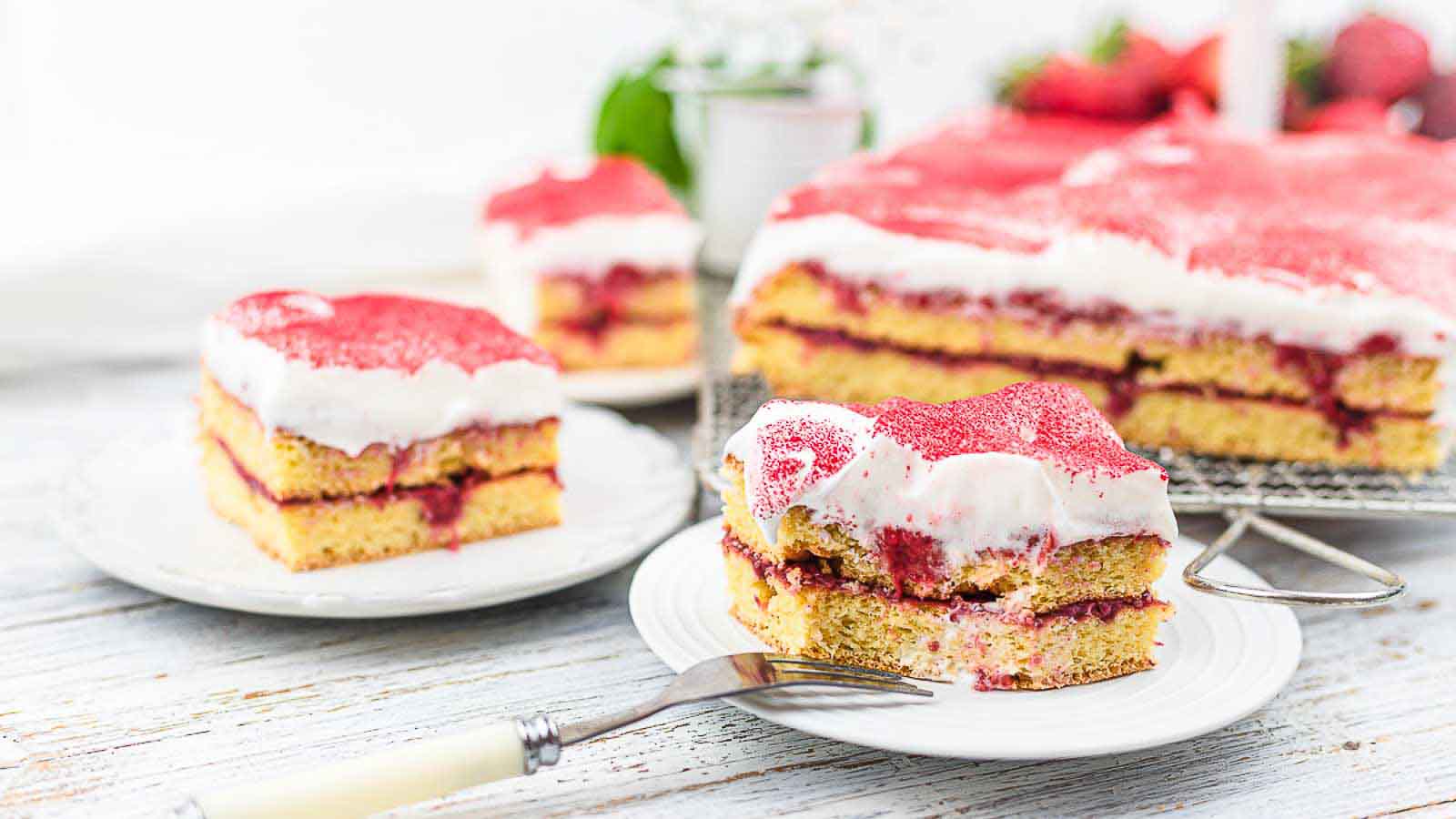 A close-up of layered sponge cake with cream and red fruit filling, topped with white frosting and red sprinkles, served on white plates.
