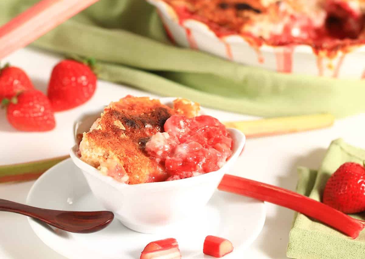 A bowl of strawberry rhubarb cobbler sits on a white plate with a spoon, surrounded by fresh strawberries and rhubarb stalks.