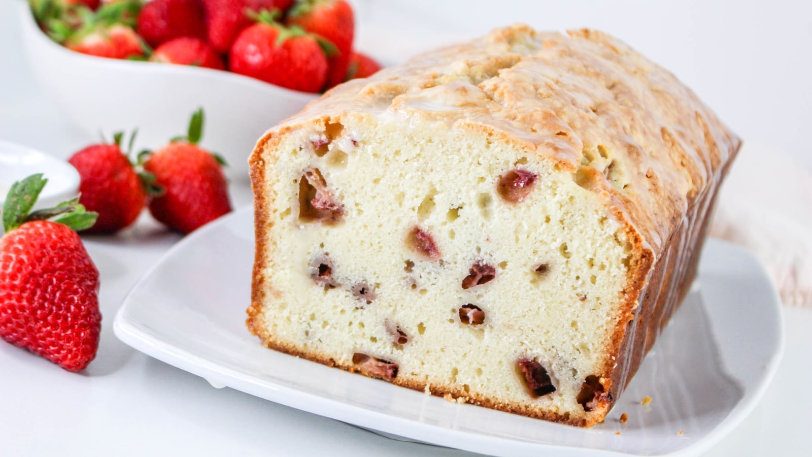A loaf of glazed strawberry bread sits on a white plate, with fresh strawberries and a bowl of strawberries in the background.