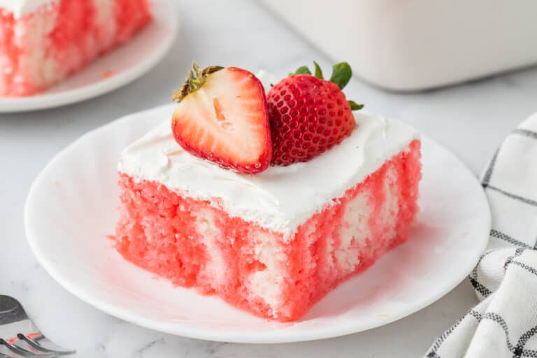 A slice of pink and white poke cake with white frosting, topped with two fresh strawberries, on a white plate.