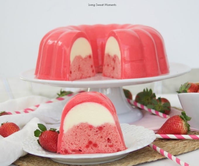 A sliced strawberry and cream gelatin dessert sits on a cake stand, with a plated slice in front. Fresh strawberries and pink striped straws are nearby.