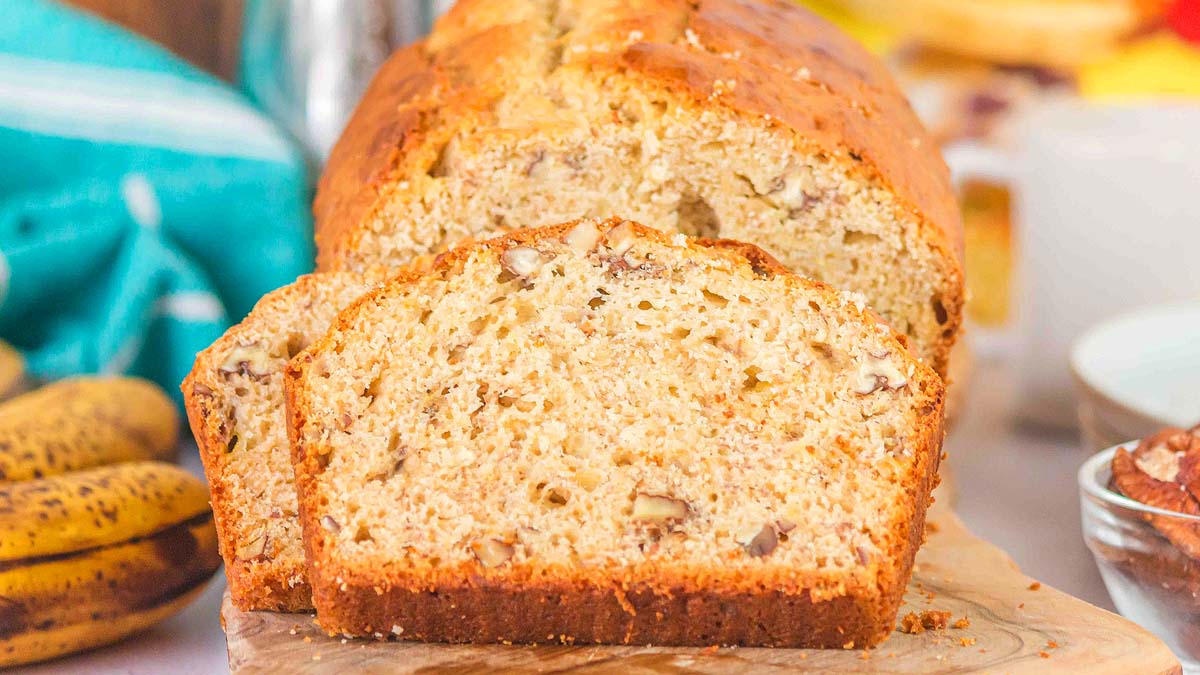 A loaf of banana bread with visible nuts, partially sliced, sits on a wooden board with bananas and a bowl in the background.