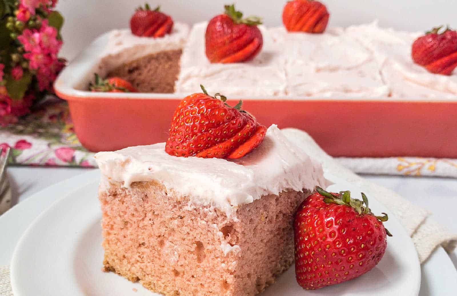 A square slice of pink frosted cake topped with strawberries sits on a white plate, with a baking dish of similar cake in the background.