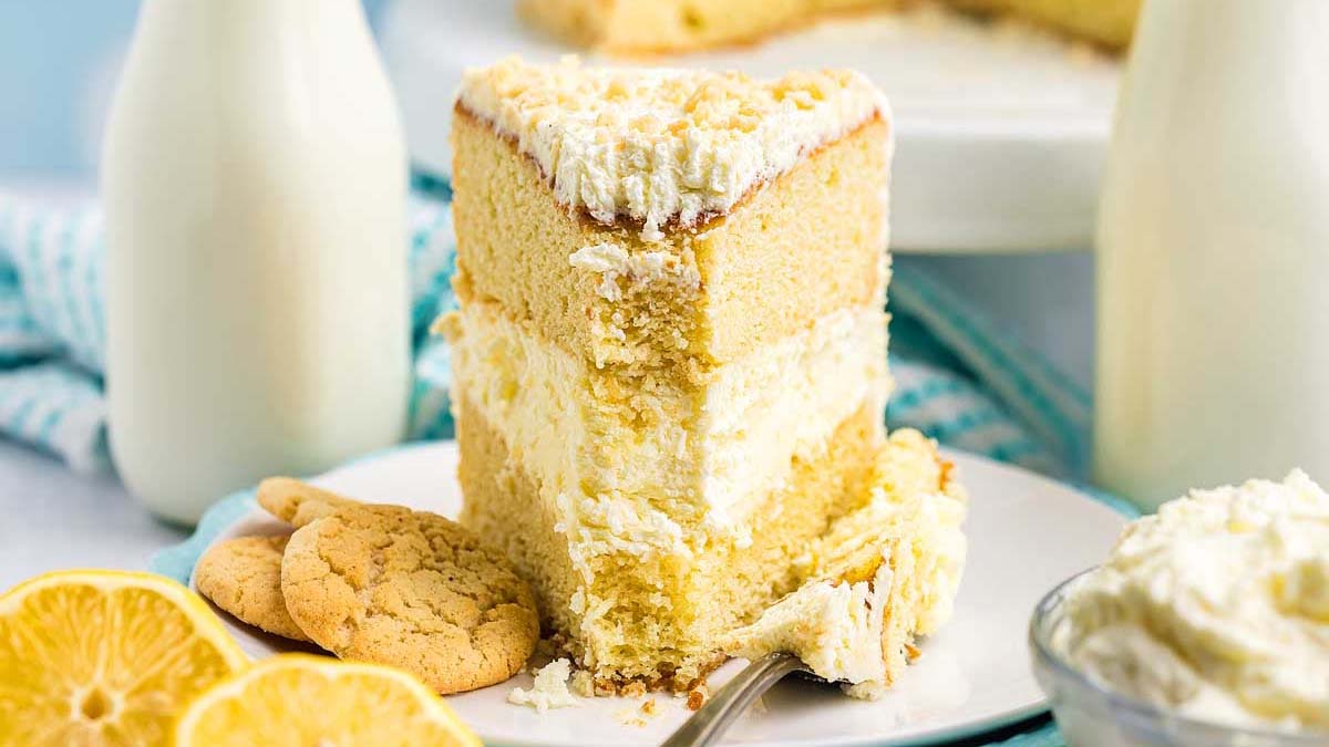 A slice of layered lemon cake with cream filling on a plate, accompanied by cookies, lemon slices, and bottles of milk.