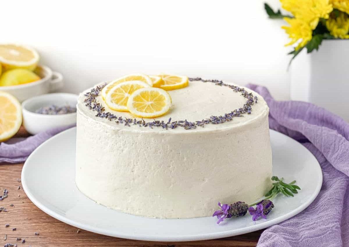 A round white frosted cake topped with lemon slices and lavender flowers sits on a white plate, with lemons and yellow flowers in the background.
