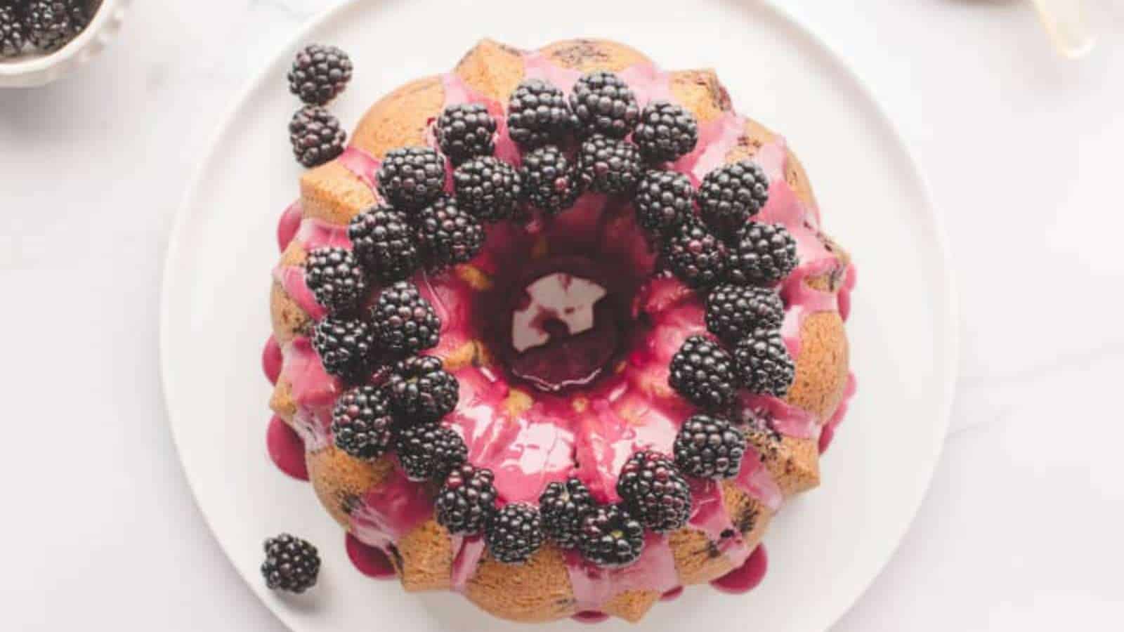 A bundt cake with pink glaze, topped with fresh blackberries, sits on a white plate.