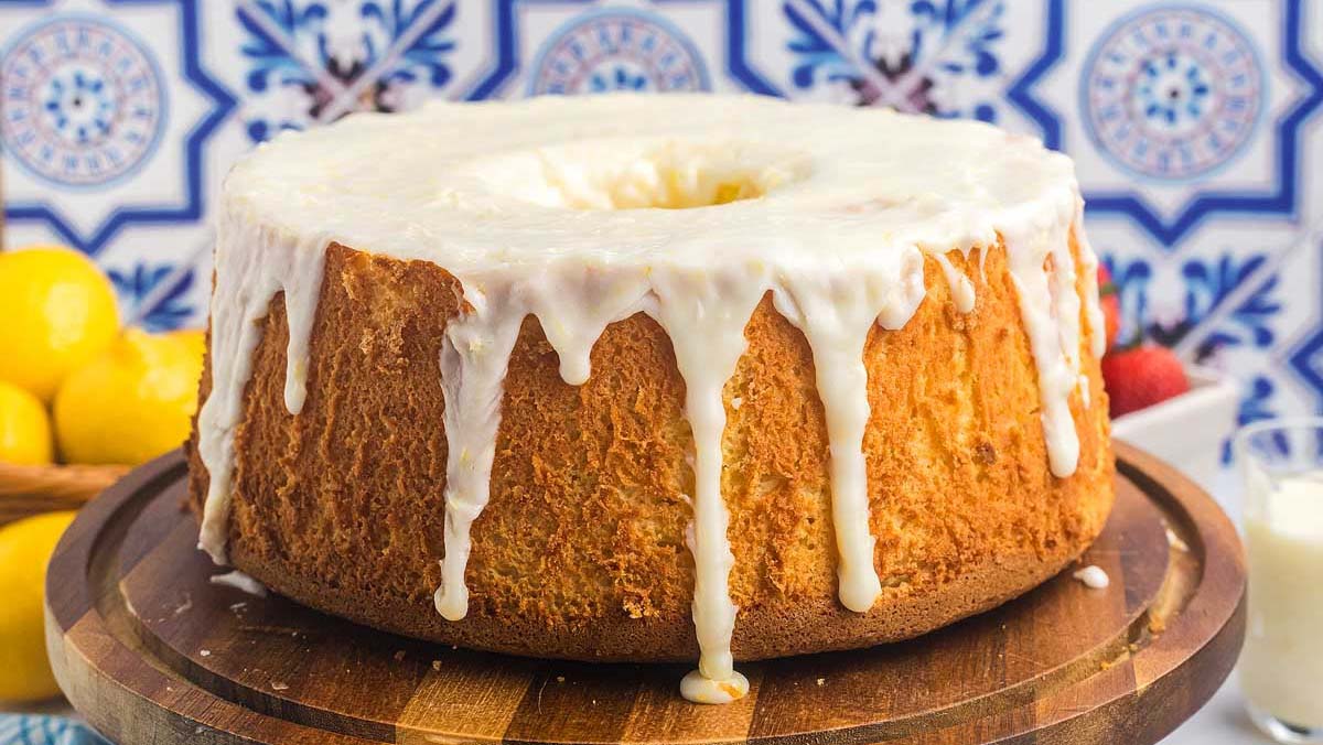 A lemon bundt cake with white icing drips sits on a wooden platter; lemons and strawberries are in the background.