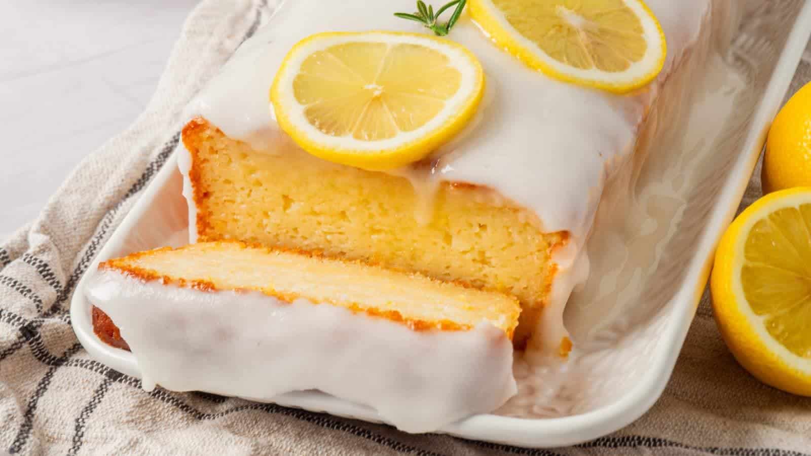 A loaf of lemon cake with white icing, topped with lemon slices, sits on a rectangular white plate with a slice cut.