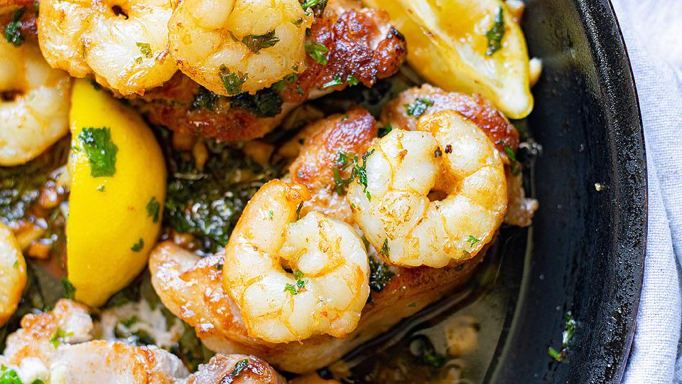 Cooked shrimp garnished with herbs, served with lemon wedges and pieces of chicken in a skillet.