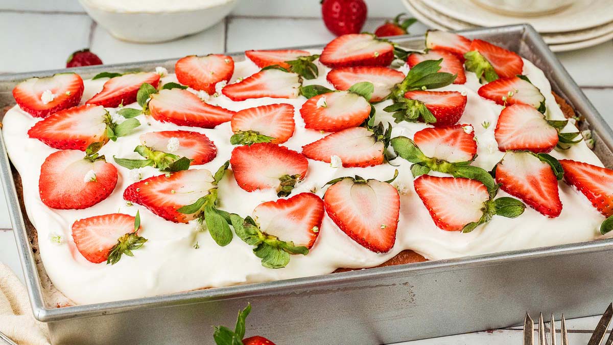 Rectangular cake topped with a thick layer of whipped cream and sliced fresh strawberries, served in a metal baking pan.
