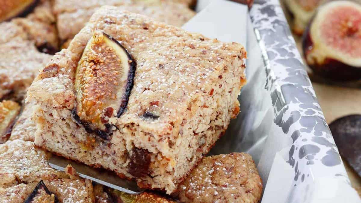 A close-up of a tray of freshly baked fig bars, with a square piece being lifted out, showing a fig slice on top and a dusting of powdered sugar.