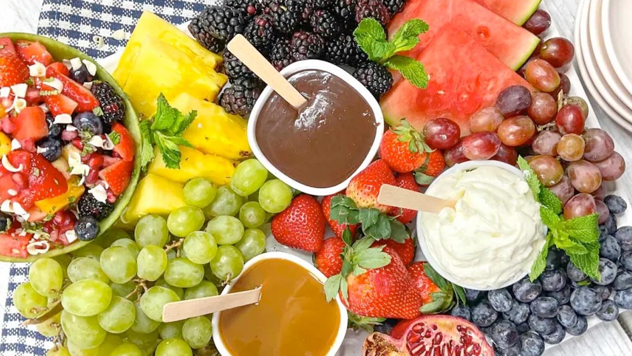 A platter with assorted fresh fruits, including grapes, strawberries, watermelon, blackberries, pineapple, and pomegranate, surrounds bowls of chocolate, caramel, and cream dips.