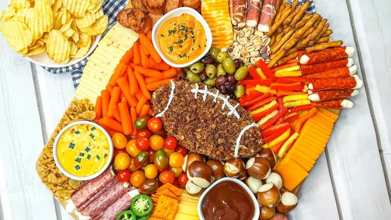 A charcuterie board with cheese, meats, crackers, dips, vegetables, and a football-shaped cheese ball, arranged for a party. A bowl of potato chips is on the side.