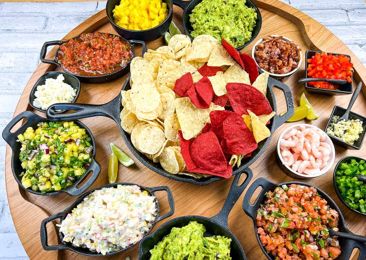 A round wooden tray with assorted dips, salsas, guacamole, shrimp, and a central plate of yellow and red tortilla chips in small black bowls.