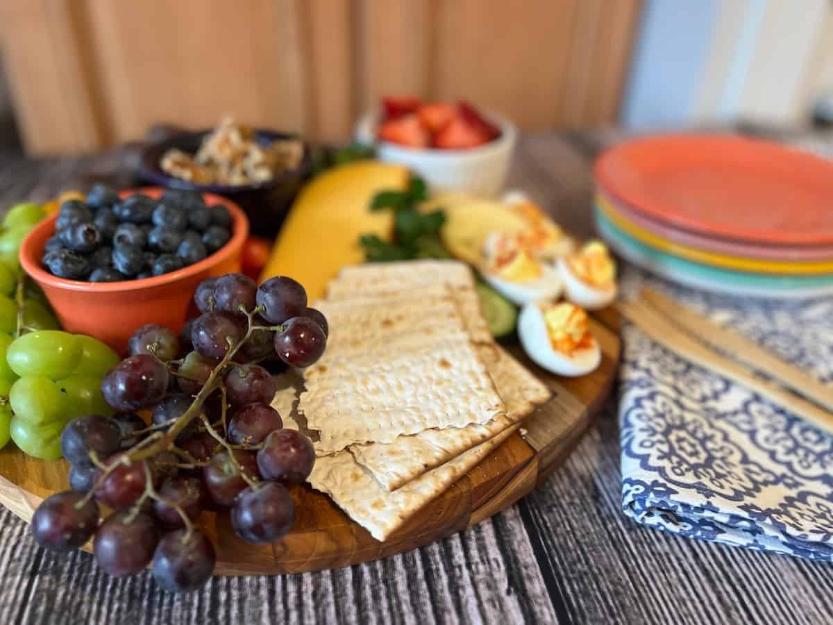 A wooden platter with matzo crackers, grapes, blueberries, deviled eggs, fruit, nuts, and cheese next to stacked colorful plates and a patterned napkin.